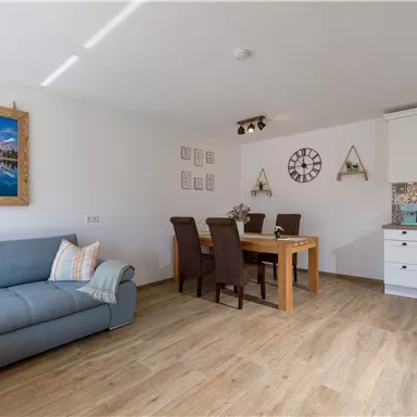 A bright living room with a blue sofa and a dining table. Pictures and a clock are hung on the wall.