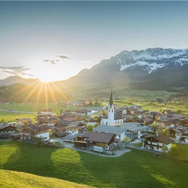 A picturesque mountain landscape with a small village and a church in the foreground. The sun rises behind the mountains, creating a wonderful atmosphere.