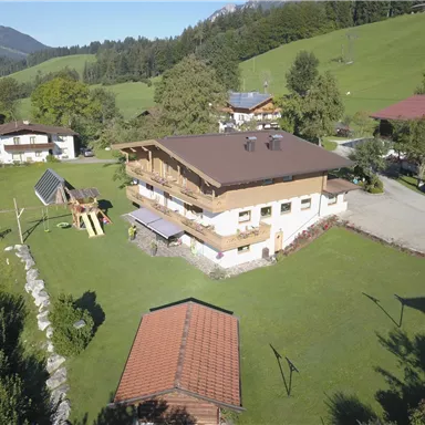 Ein modernes, zweistöckiges Haus umgeben von grünem Rasen und Bäumen. Im Vordergrund befindet sich ein Spielplatz und im Hintergrund Hügel und Berge.