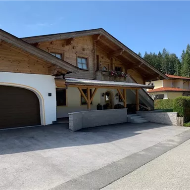 A cozy house in the so-called Alpine style with wooden elements. The entrance area is designed in a friendly way and there is a paved driveway.