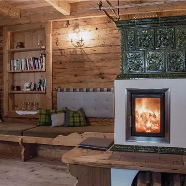 A cozy room with wooden walls and a classic tiled stove. A seating area with cushions invites you to relax.