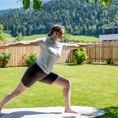 A woman is doing yoga on a mat outdoors. In the background, green meadows and mountains can be seen.