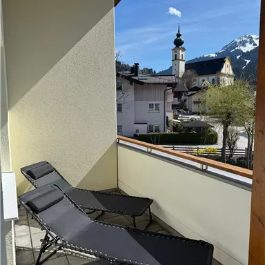 A balcony with two loungers and a beautiful view of a small village. In the background, the mountains and a church tower are visible.