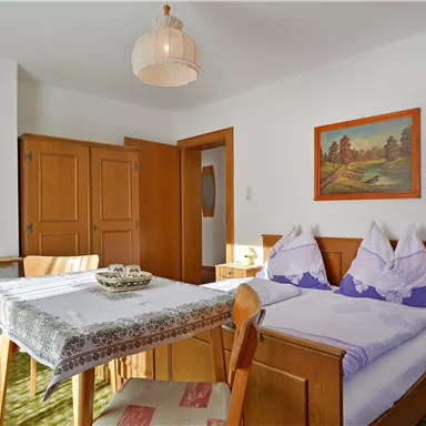 A cozy room with two beds, a table, and wooden furniture. The walls are light-colored, and there is a picture as well as a lamp on the nightstand.
