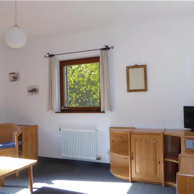 Ein helles Wohnzimmer mit Holzmöbeln und einem Fenster, das Licht hereinlässt. Es gibt einen Tisch, Stühle und einen Fernseher.