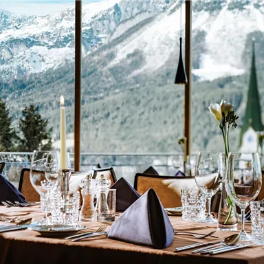 An elegant table set with dishes and glasses, beautifully arranged for a meal. In the background, snow-covered mountains and a green church tower can be seen.