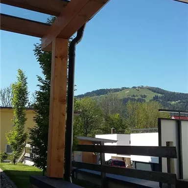 Eine Aussicht auf einen grünen Hügel unter klarem blauen Himmel. Im Vordergrund ist eine Holzstruktur und einige Bäume sichtbar.