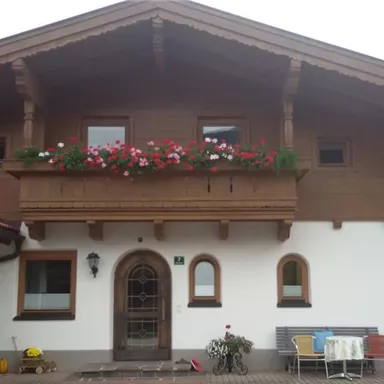 Ein traditionelles Haus mit Holzverkleidung und einem Balkon voller Blumen. Der Eingangsbereich ist einladend mit einem Tisch und Stühlen vor der Haustür.