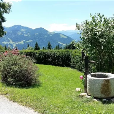 A picturesque landscape with a well-maintained garden and a fountain pedestal. In the background, gentle hills and mountains can be seen under a clear sky.