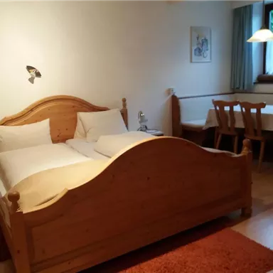 A cozy room with a wooden double bed and two pillows. There is a table with chairs and a window with a view outside.