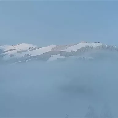 A snowy landscape with hills partially shrouded in mist. The sky is clear and blue.