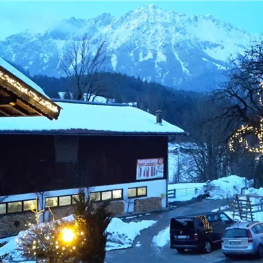 A snowy landscape with mountains in the background and a cozy building. The evening sky is blue and warmly illuminated by lights on a tree.
