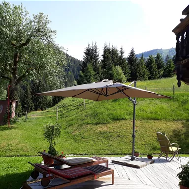 Ein schöner Garten mit einer Terrasse, Liegen und einem Sonnenschirm. Im Hintergrund sind grüne Wiesen und Bäume zu sehen.