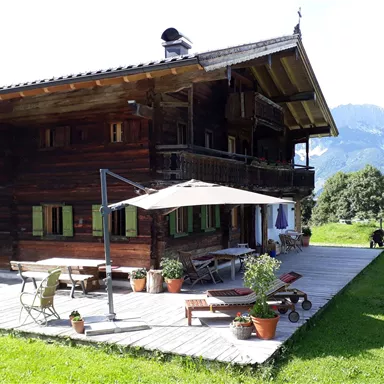 Ein rustikales Holzhaus mit einem großen Deck und mehreren Töpfen mit Pflanzen. Im Hintergrund sind sanfte Hügel und Berge sichtbar.