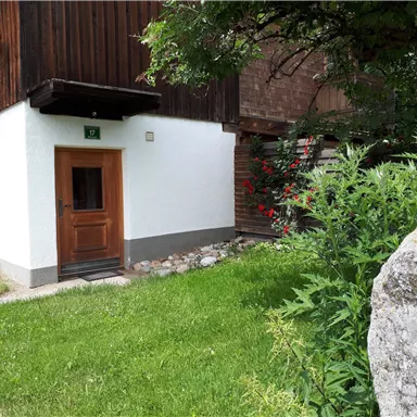 Ein gemütliches Haus mit einer weißen Fassade und einer Holzveranda. Es umgibt grünes Gras und bunte Blumen.