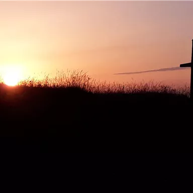 A sunset over a hilltop with tall grass. In the foreground, a cross is silhouetted against the colorful sky.