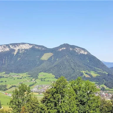 An impressive mountain landscape with gentle hills and a clear blue sky. In the foreground, lush trees and a small village are visible.