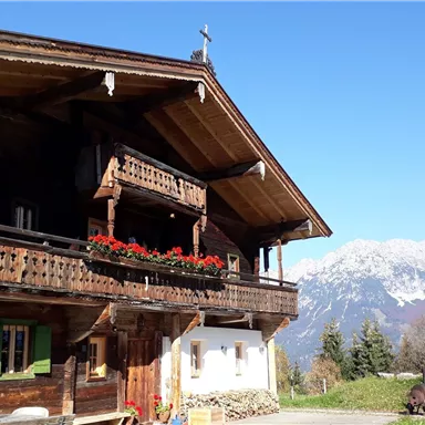 Ein traditionelles Holzhaus mit Balkonen und bunten Blumenkästen. Im Hintergrund sind majestätische Berge und ein blauer Himmel zu sehen.