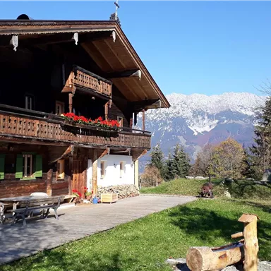 Ein traditionelles Holzhaus mit Balkon und bunten Blumen. Im Hintergrund sind schneebedeckte Berge und ein klarer blauer Himmel zu sehen.
