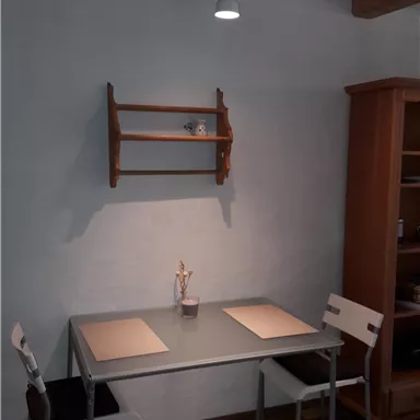 Ein einfaches Esszimmer mit einem Tisch und zwei Stühlen. An der Wand befindet sich ein kleiner Holzregal.