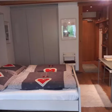 Ein gemütliches Schlafzimmer mit einem Doppelbett und einer neutralen Farbpalette. Im Raum befinden sich ein Tisch und Regale, die zusätzliche Funktionalität bieten.