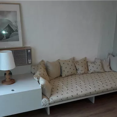 Ein gemütliches Wohnzimmer mit einem langen Sofa, das mit Kissen dekoriert ist. Neben dem Sofa befindet sich ein Beistelltisch mit einer Lampe und einem Klimagerät.