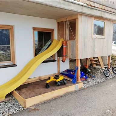 A play area with a yellow slide and a playhouse. Additionally, toys such as a Bobbycar and shovels can be seen.