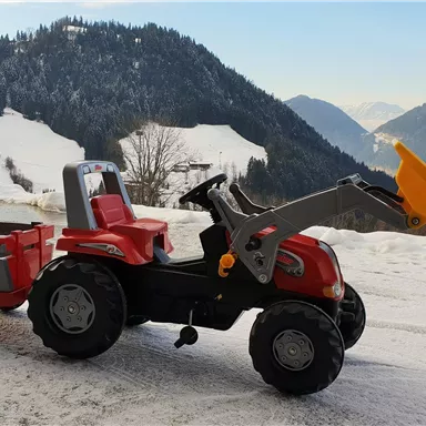 A red toy tractor with a trailer is on a snowy terrain. In the background, there are mountains and a blue sky.