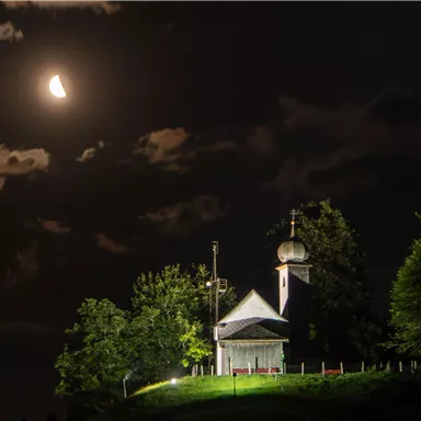A small chapel stands on a hill under a clear night sky. The moon shines and clouds drift by.