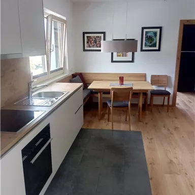 A modern kitchen with bright furnishings and a cozy dining area. The walls are decorated with artwork, and plenty of natural light floods through the window.