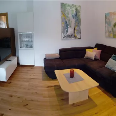 A cozy living room with a large couch and a coffee table. Modern artworks hang on the walls, and the floor is made of light wood.