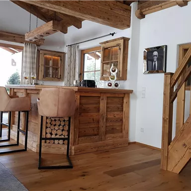 Ein gemütlicher Raum mit Holzmöbeln und einer offen gestalteten Bar. Große Fenster sorgen für viel Tageslicht und eine einladende Atmosphäre.