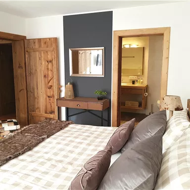 Ein gemütliches Schlafzimmer mit Holzdetails und einem großen Bett. Im Hintergrund sind ein Spiegel und ein Badezimmer sichtbar.