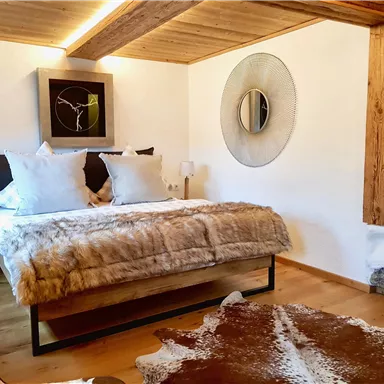 Ein stilvolles Schlafzimmer mit einem großen Bett, dekoriert mit kuscheligen Fellen. An den Wänden sind moderne Kunstwerke und ein runder Spiegel zu sehen.