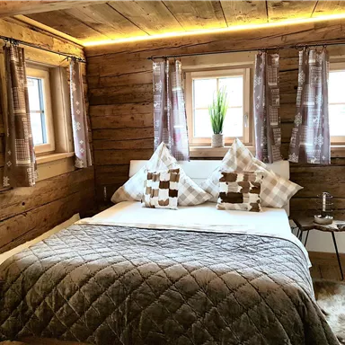 Ein rustikales Schlafzimmer mit Holzverkleidung und zwei Fenstern. Das Bett ist gemütlich dekoriert mit Kissen und einer warmen Decke.