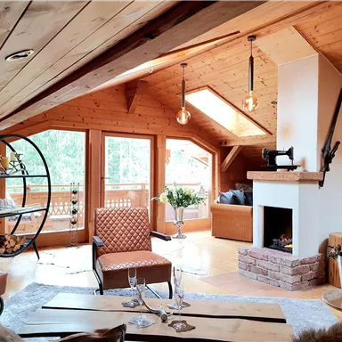 Ein gemütliches, rustikales Wohnzimmer mit Holzwänden und großen Fenstern. Im Raum befinden sich komfortable Möbel und ein Kamin, der eine einladende Atmosphäre schafft.