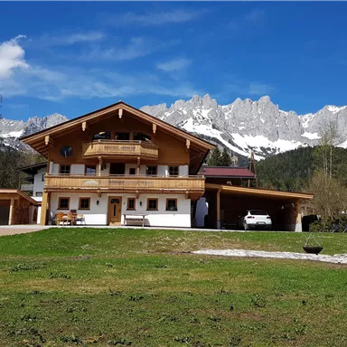 Ein schönes Holzhaus mit einem großen Garten. Im Hintergrund sind majestätische Berge und ein klarer blauer Himmel zu sehen.