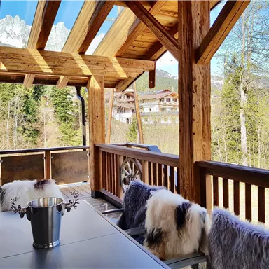Eine gemütliche Veranda mit Holzmöbeln und warmen Fellen. Im Hintergrund sind grüne Bäume und Berge zu sehen.