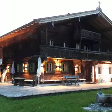 Ein traditionelles Holzhaus in den Bergen, beleuchtet von warmen Lichtern. Im Hintergrund sind die Berge und die natürliche Landschaft zu sehen.