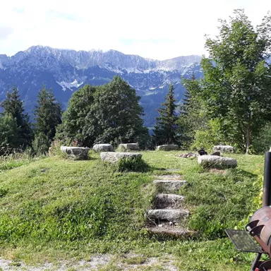 Eine malerische Landschaft mit Blick auf hohe Berge und einer Wiese. Im Vordergrund sind Steinquader und einige Bäume zu sehen.