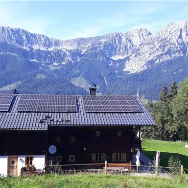 Ein traditionelles Haus mit Solarpanelen, umgeben von grünen Wiesen. Im Hintergrund erheben sich majestätische Berge unter blauem Himmel.