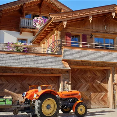 Ein traditionelles Holzhaus mit einem schönen Balkon und bunten Blumen. Vor dem Haus steht ein orangefarbener Traktor neben einem grünen Anhänger.