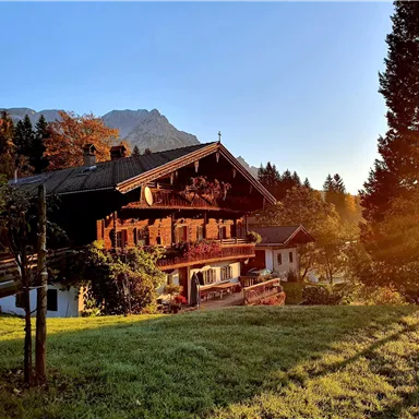 A traditional alpine house surrounded by green meadows and trees. The sun rises and bathes the scene in warm light.