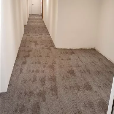 Ein langer, heller Gang mit grauem Teppichboden. An den Wänden befinden sich weiße Wände und einige Deckenlichter.