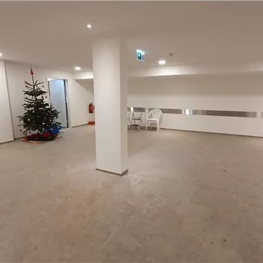 Ein leerer Raum mit hellem, neutralem Design. In der Ecke steht ein Weihnachtsbaum und einige Stühle sind vorhanden.