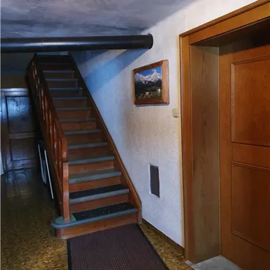 An inviting hallway with a wooden staircase and a painted landscape picture on the wall. The walls are light and the floor is covered with a carpet.