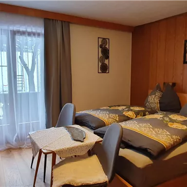 A cozy bedroom with two beds, a small table, and a view of the balcony. The walls are made of wood and there are curtains that let in light.