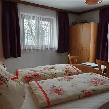 A cozy bedroom with a double bed and colorful pillows. Large windows let in plenty of light, and there is a wooden wardrobe and a chair in the room.