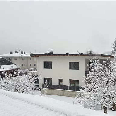 Ein verschneites Wohngebiet mit weißen Gebäuden und Bäumen. Der Himmel ist grau und die Landschaft ist winterlich ruhig.