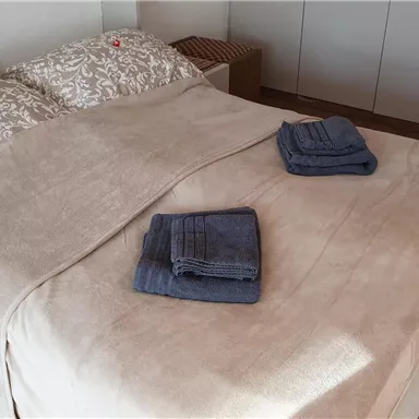Ein gemütliches Schlafzimmer mit einem Bett und sauberer Bettwäsche. Auf der Decke liegen zwei gefaltete Handtücher.